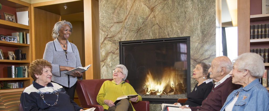Group of seniors sitting by fireplace discussing a book.