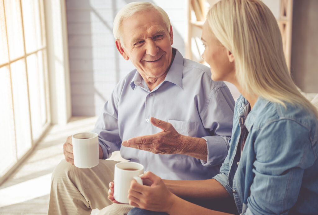 An elderly man and his daughter sit and chat in a sunny room with cups of coffee.