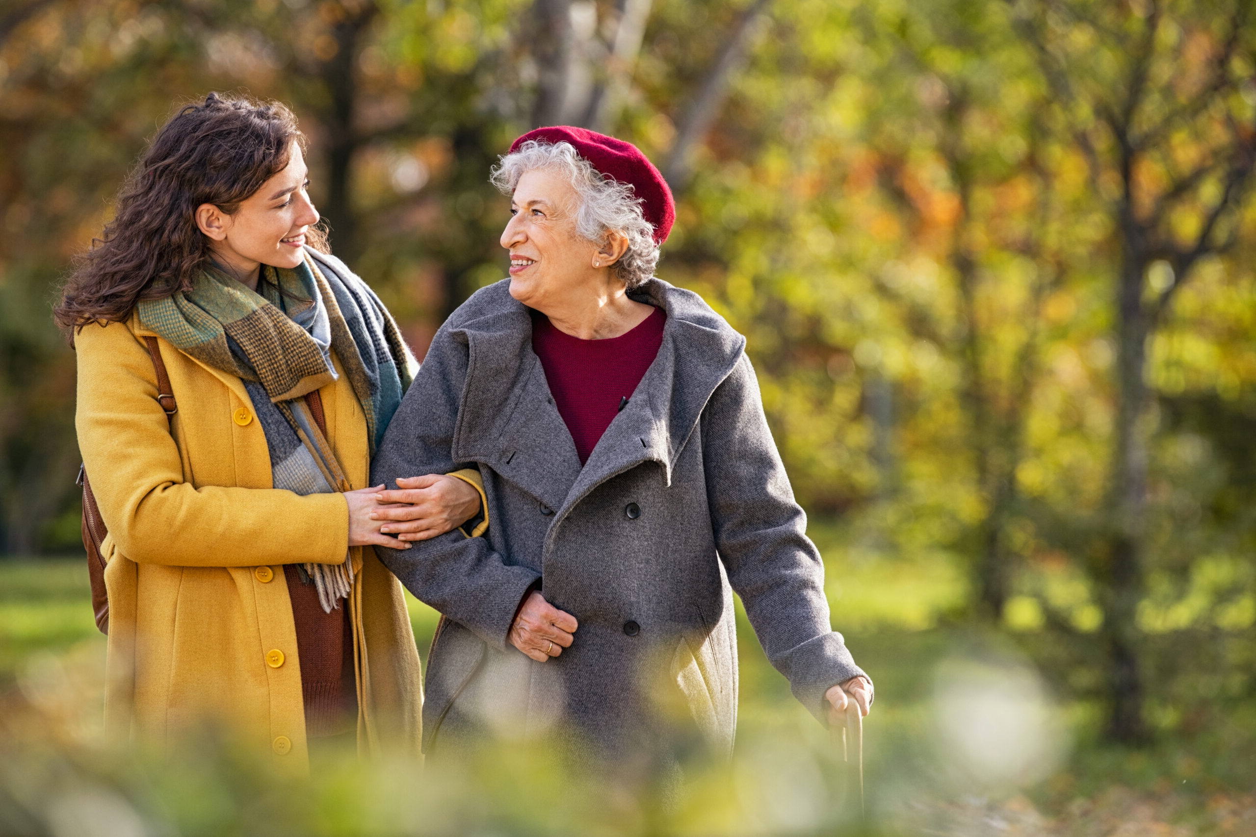 Young woman in park wearing winter clothing walking with old grandmother. Happy grandma wearing coat walking with lovely girl outdoor with copy space. Smiling lovely caregiver and senior lady walking in park during autumn and looking at each other.